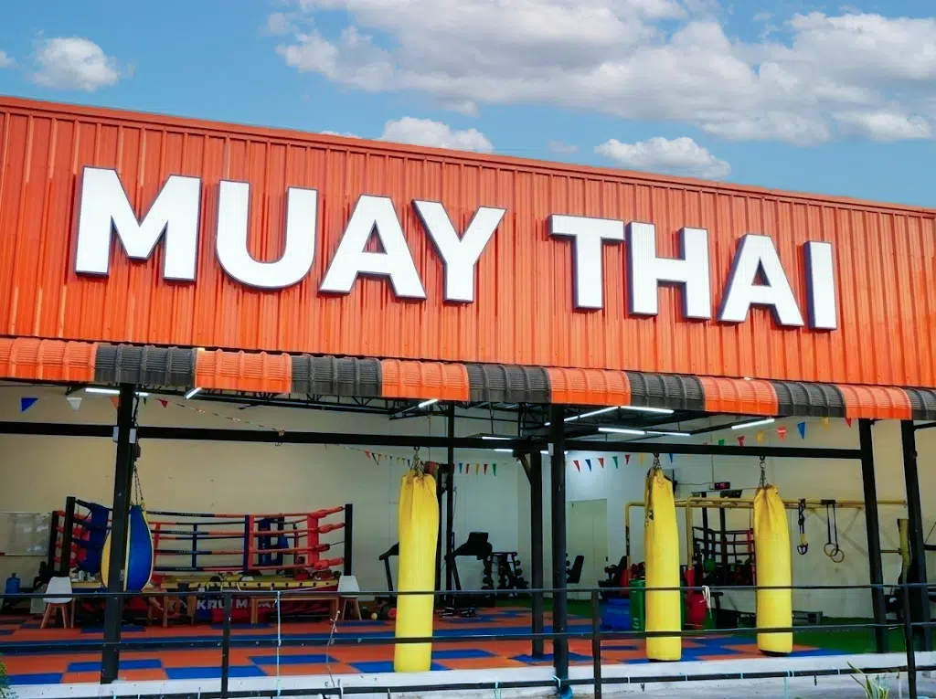 ป้ายหน้าค่ายมวยไทย "บ้านครูมวย" ตัวอักษร MUAY THAI ขนาดใหญ่ เห็นบรรยากาศยิมที่กว้างขวางและอุปกรณ์กระสอบทรายครบครัน